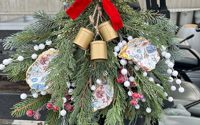 A festive holiday decoration with greenery, a red bow, golden bells, and painted ornaments, hangs on a vehicle.