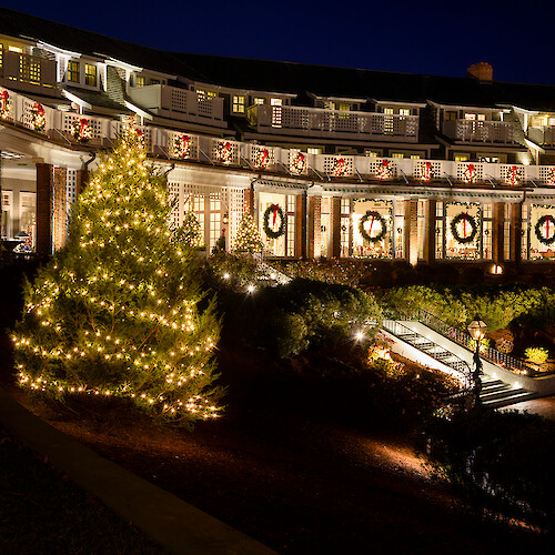 A festively lit building with wreaths and ribbons, a glowing Christmas tree, and a stairway with lights, under a dark evening sky.