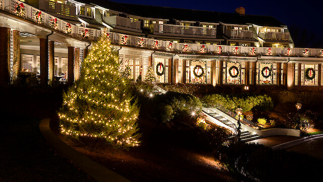 A festively lit building with wreaths and ribbons, a glowing Christmas tree, and a stairway with lights, under a dark evening sky.
