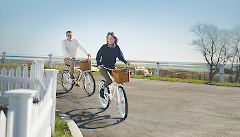Two people are enjoying a bike ride on a sunny day near a white fence and the ocean, with wicker baskets on their bikes.