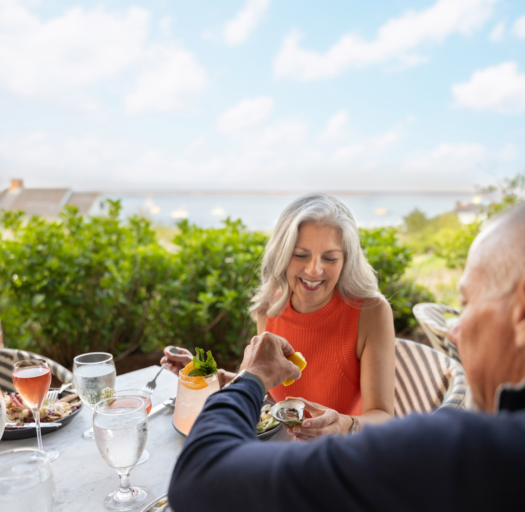 A woman in a red top enjoys a meal outdoors with a view, while a person squeezes lemon on her dish.