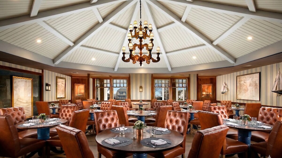A cozy restaurant with tufted leather chairs, round tables, a central chandelier, and nautical decor featuring model ships and maps.