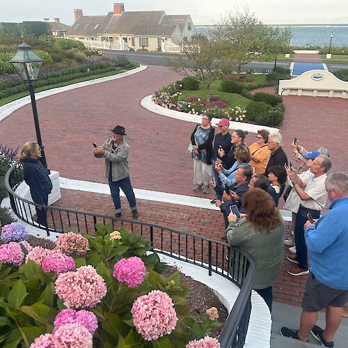 A group of people are gathered on a brick walkway, taking photos of a scene, with colorful hydrangeas in the foreground.
