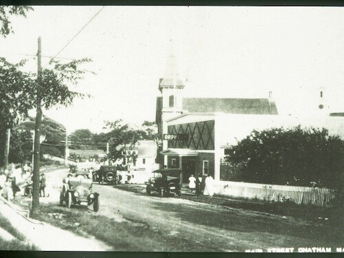 A vintage street scene with cars, people, and a building marked "Orpheum" in a small town setting, possibly early 20th century.