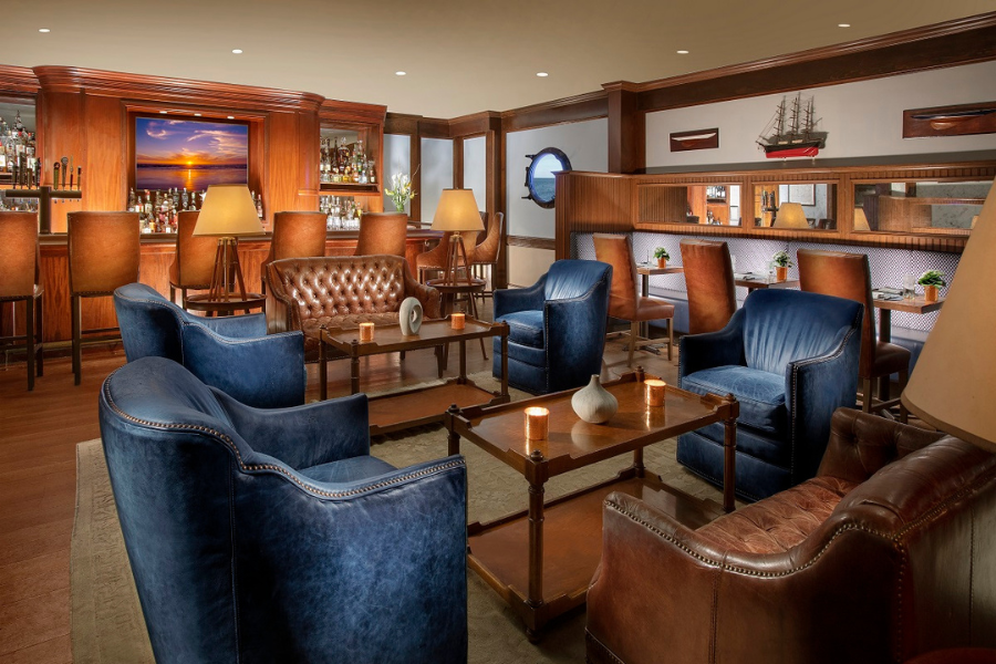 A cozy lounge with leather chairs, small tables, a bar with stools, and decorative elements like lamps and a ship model adorn the room.