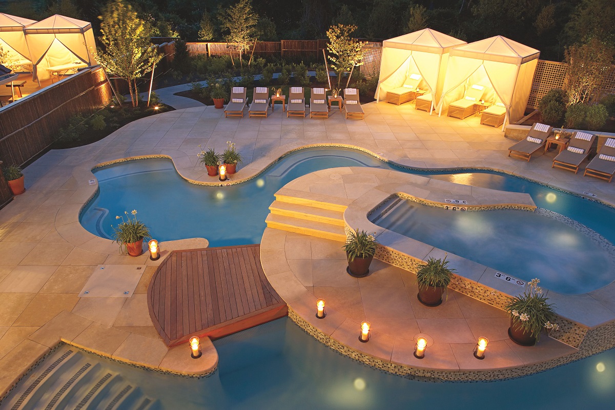 The image features a stylish outdoor pool area with loungers, cabanas, decorative lighting, and surrounding greenery.