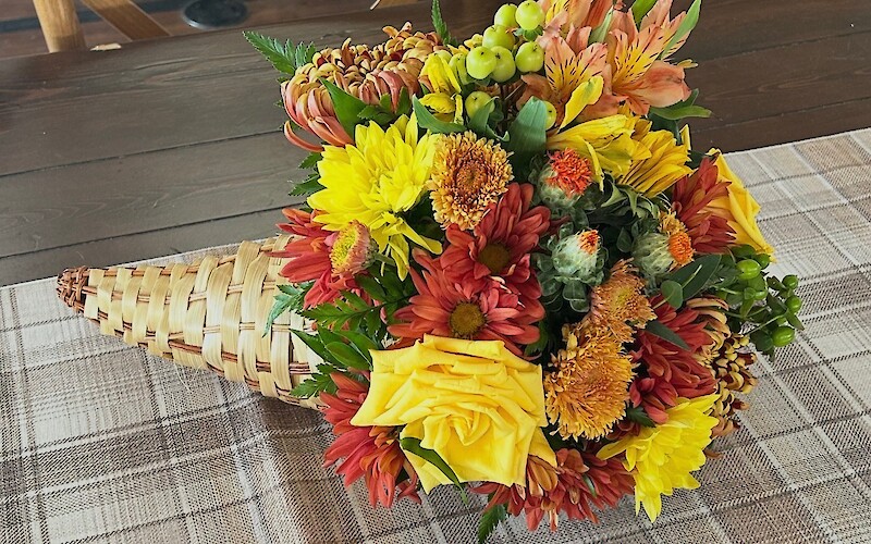 A cornucopia-style arrangement of colorful flowers on a checkered table runner and wooden table, with wooden chairs in the background.