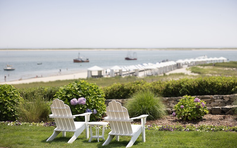 Two white chairs overlook a scenic beach with boats, cabanas, and lush gardens adorned with hydrangeas and green grass.