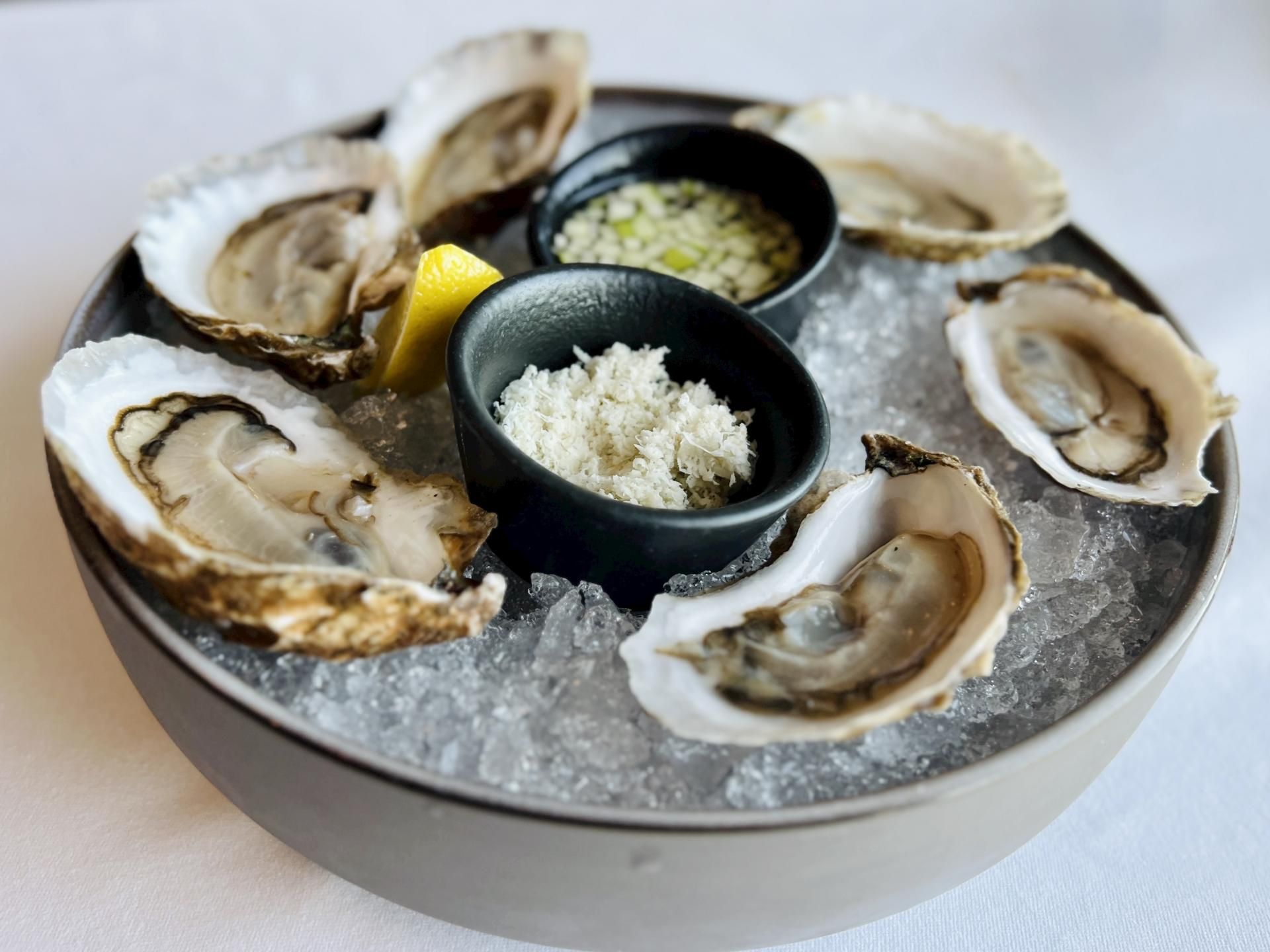 A plate of oysters on ice with lemon, accompanied by small bowls of minced garnish and grated horseradish.