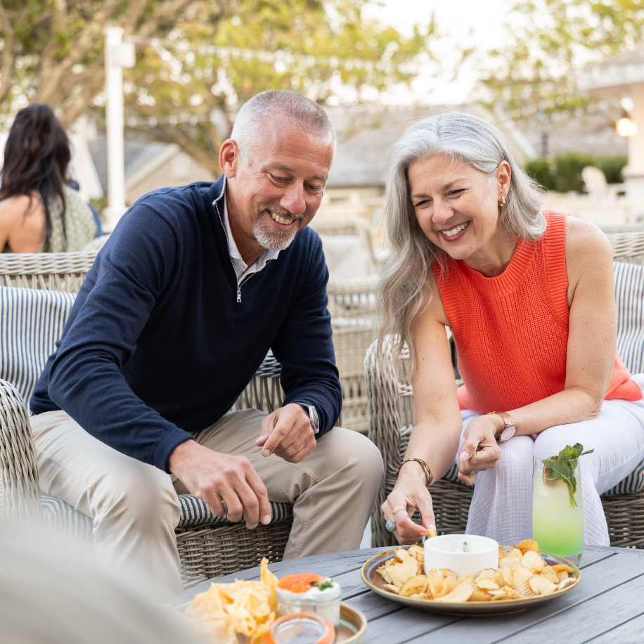 A smiling couple enjoying snacks and a drink outside, sitting on wicker chairs, with chips, dips, and a mint beverage on the table.