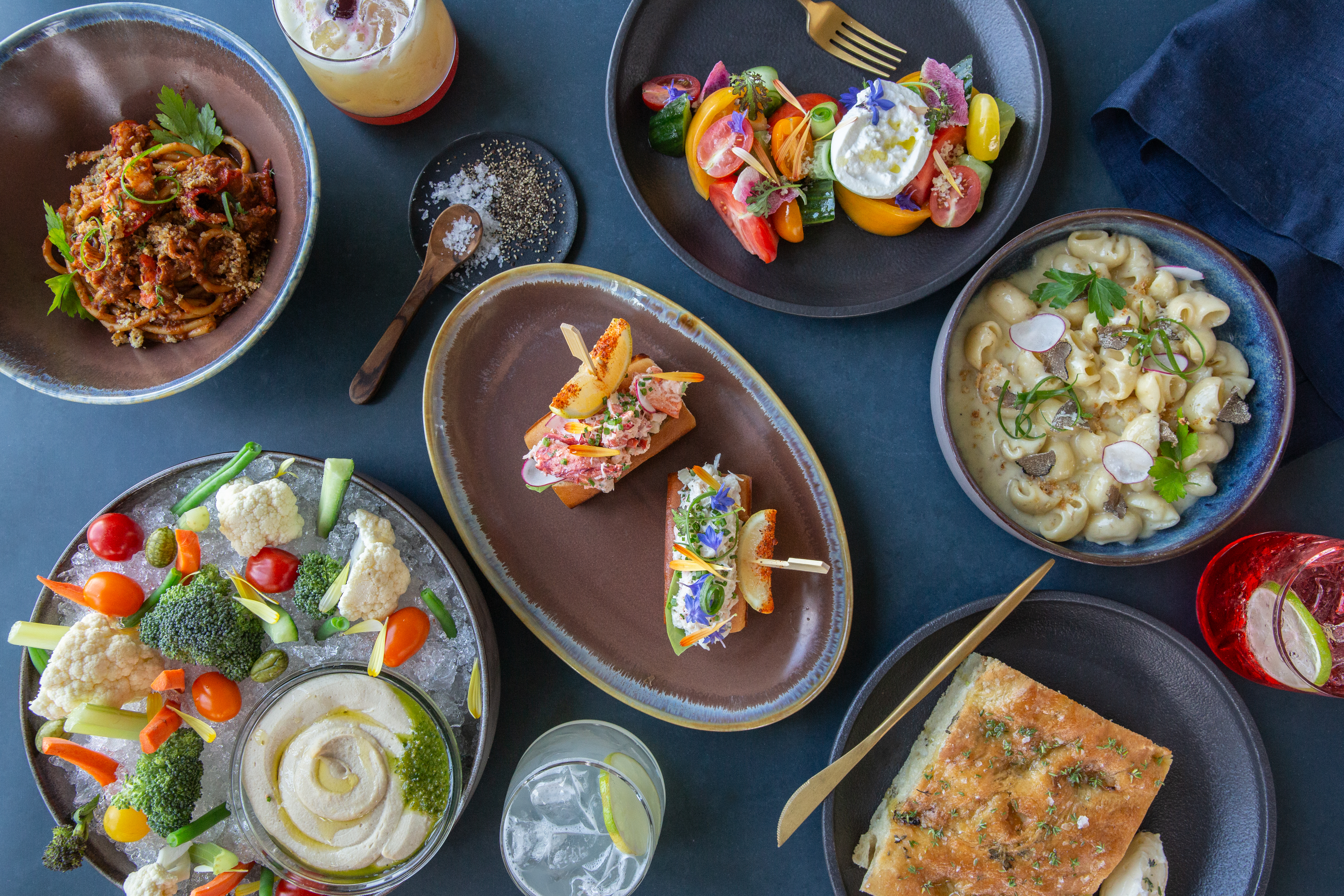 A variety of dishes including pasta, salad, lobster rolls, hummus with veggies, focaccia, and drinks.