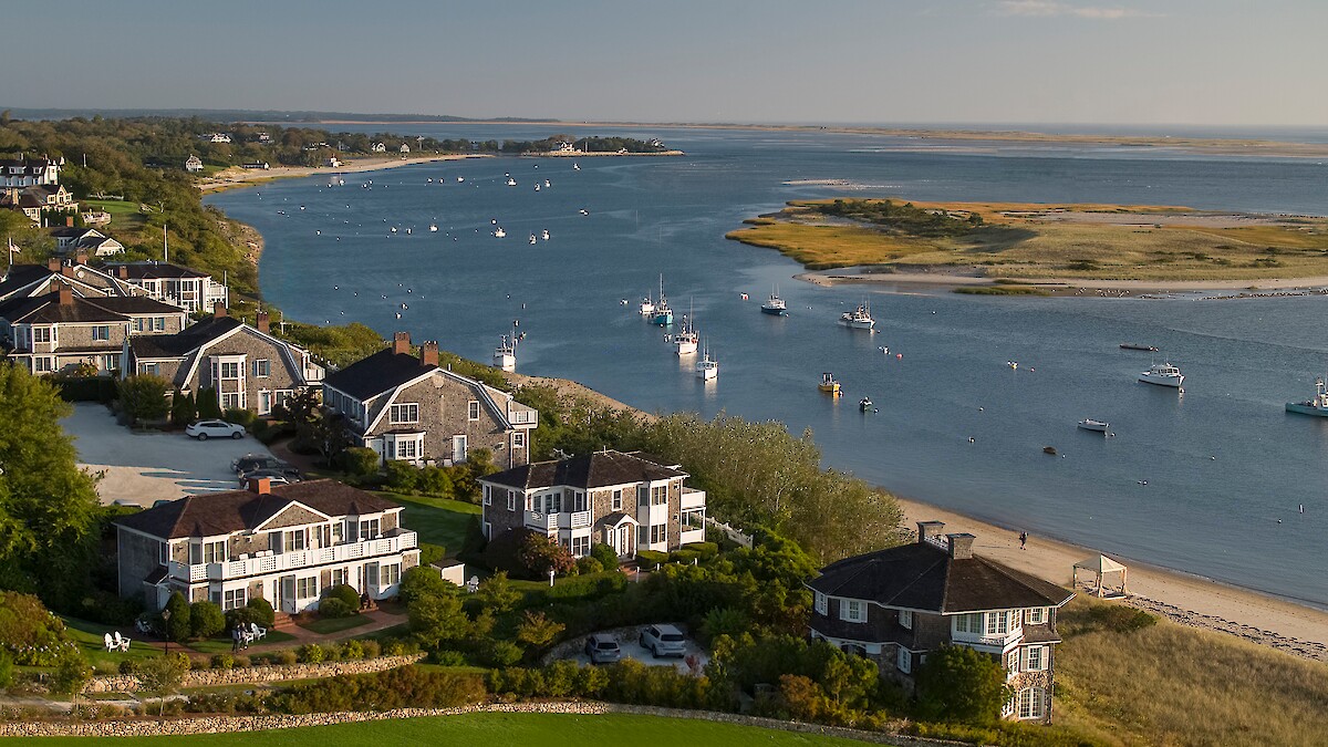 Coastal scene with waterfront homes, lush greenery, and boats on a calm blue waterway under a clear sky.