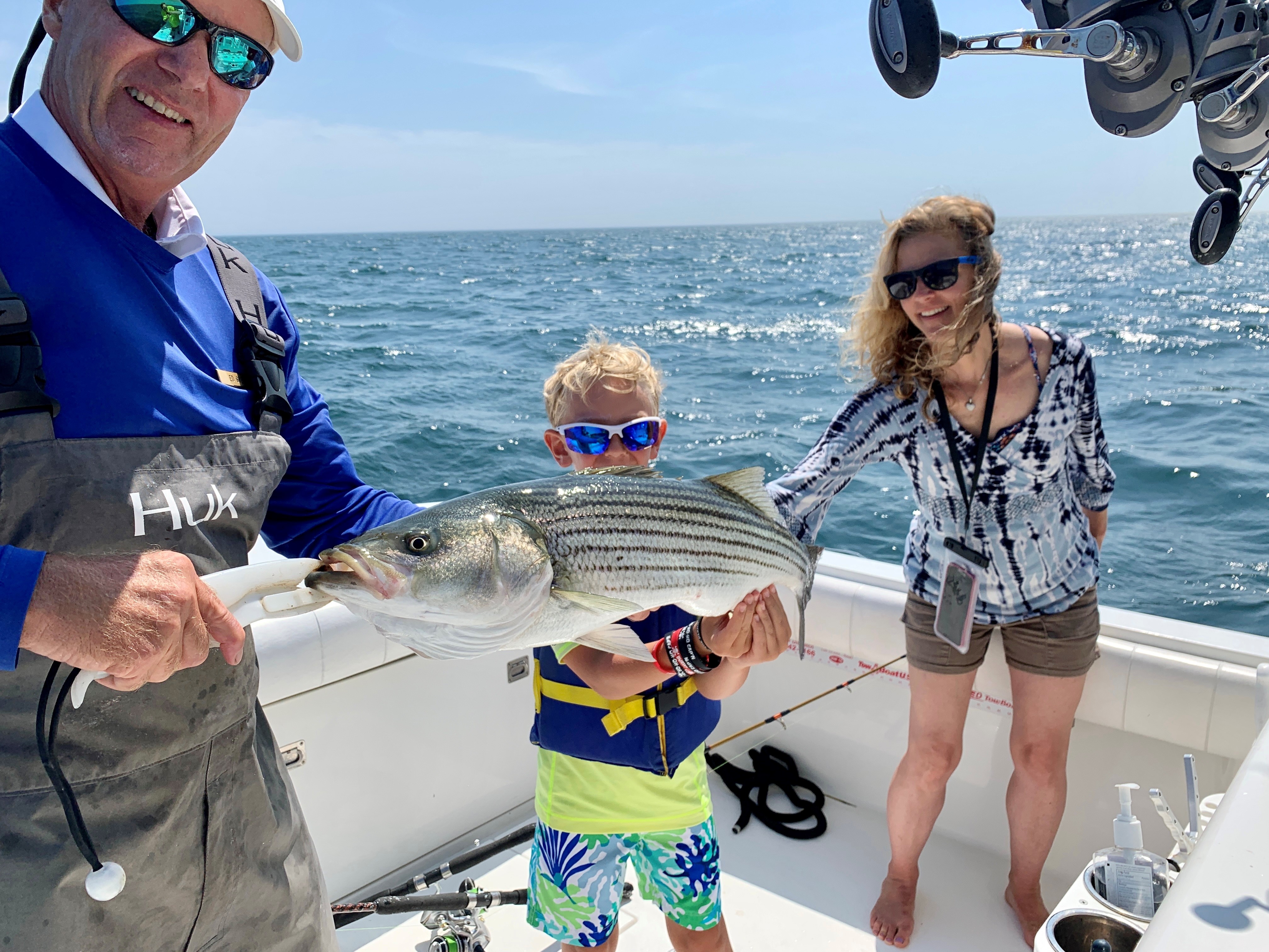 A group on a boat holds a large striped fish they've caught, with the ocean in the background, under a clear blue sky.