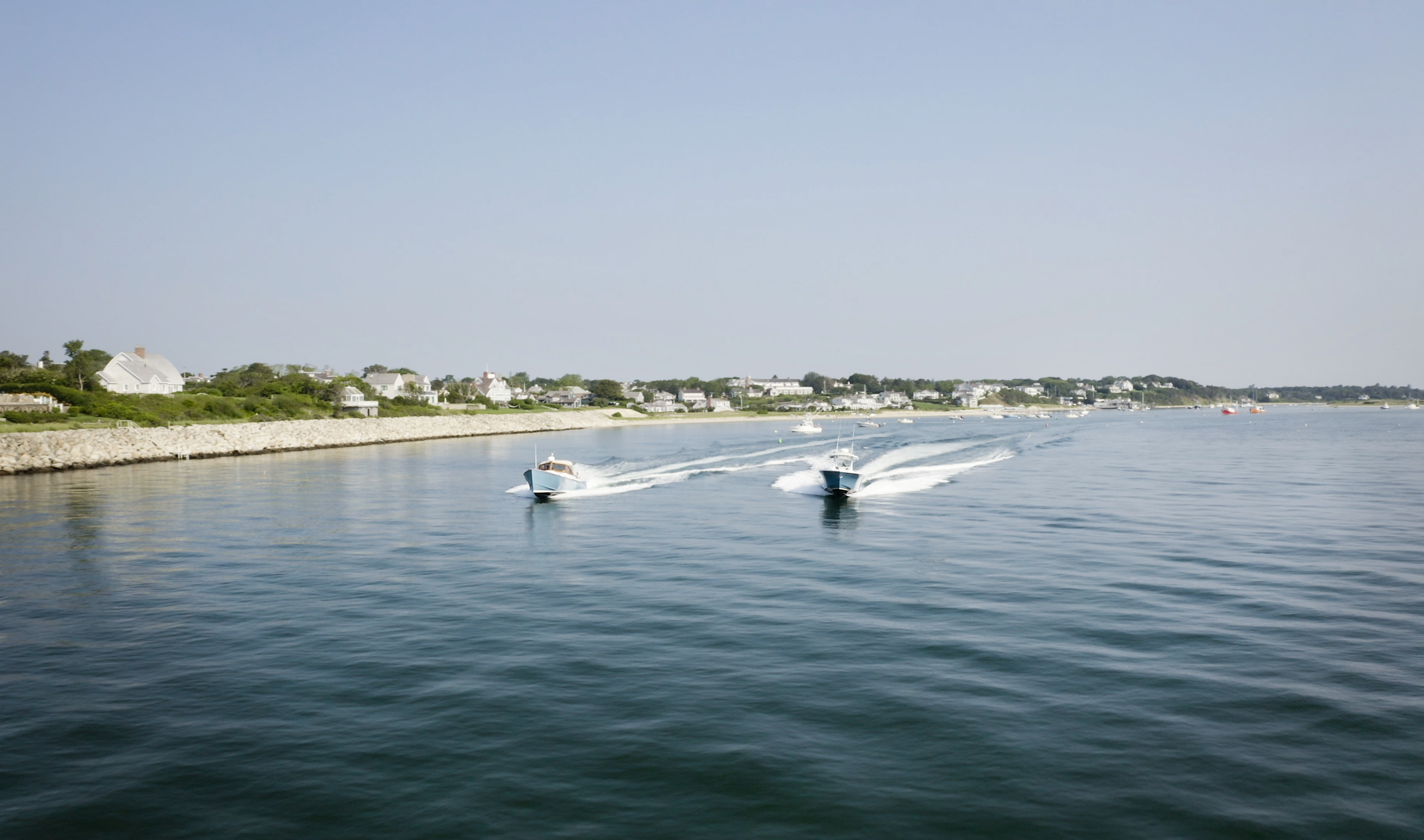 Two speedboats create waves as they move across a calm body of water near a rocky shoreline with houses in the background.