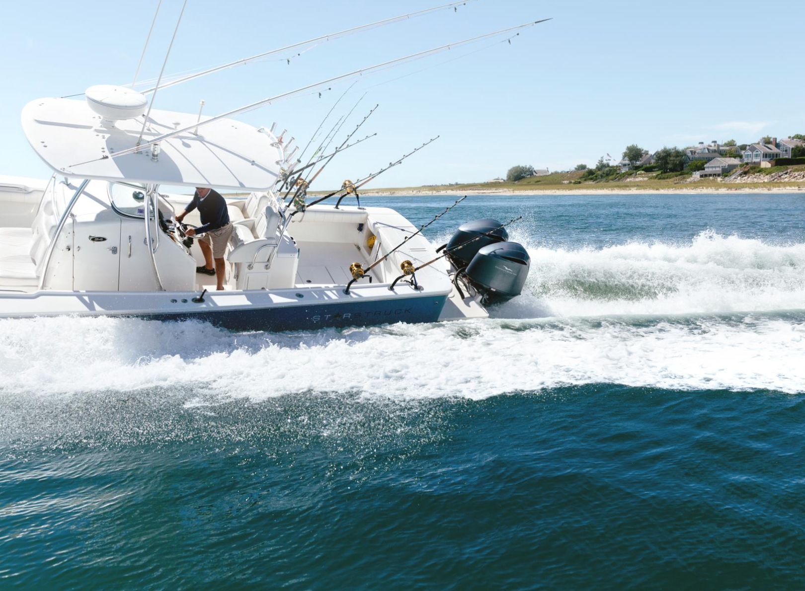 A motorboat speeds through water, with fishing rods and two outboard engines, near a coastal area with houses visible in the background.