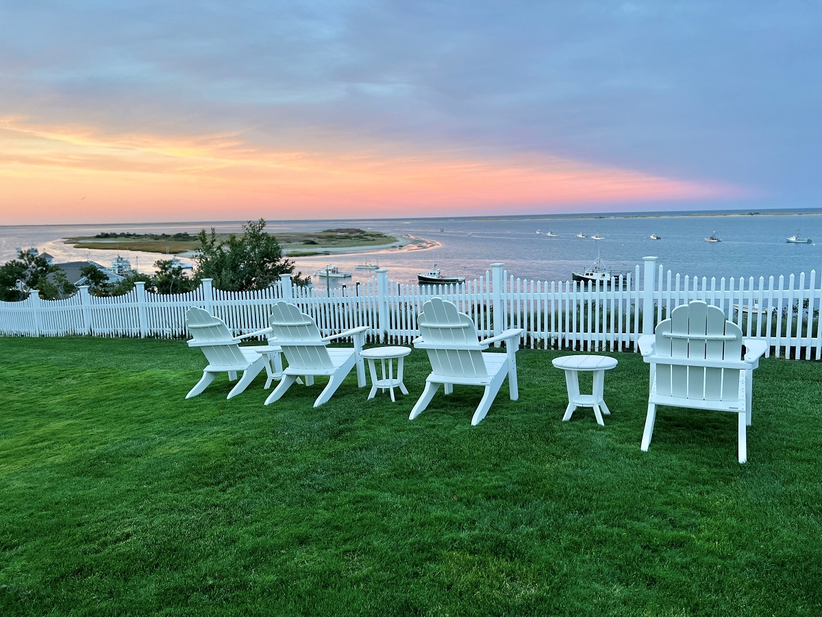 White Adirondack chairs and tables on a grassy lawn by a white fence overlook a bay with boats at sunset.