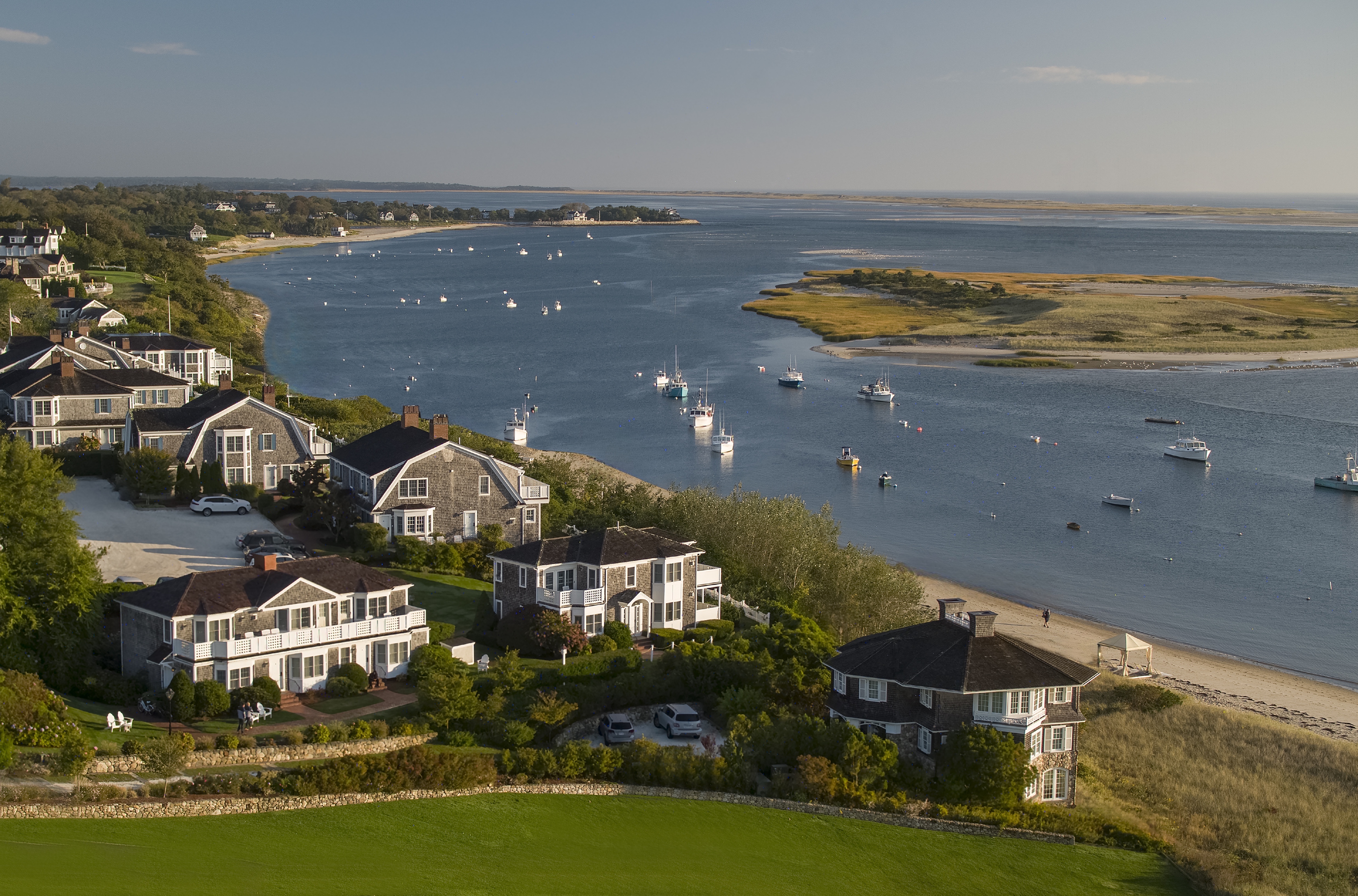 Coastal homes overlook a scenic bay with boats and sandy beaches, surrounded by green landscapes and clear blue water.