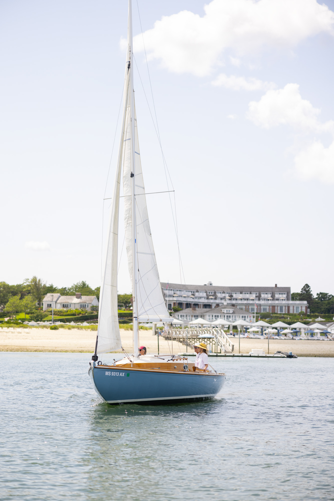 A sailboat is gliding on the water near the shore with a house and a dock visible in the background under a partly cloudy sky.