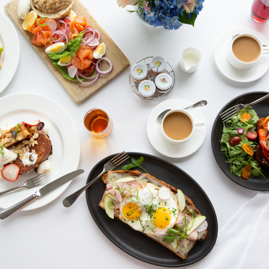 A brunch setting with open-faced sandwiches, salads, coffee, orange juice, and pastries on a white tablecloth, adorned with flowers.