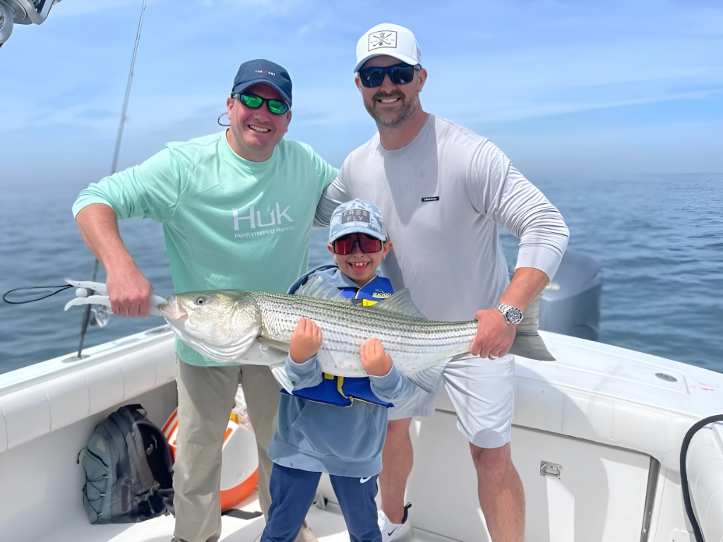 Three people on a boat proudly holding a large fish, smiling under a clear sky.