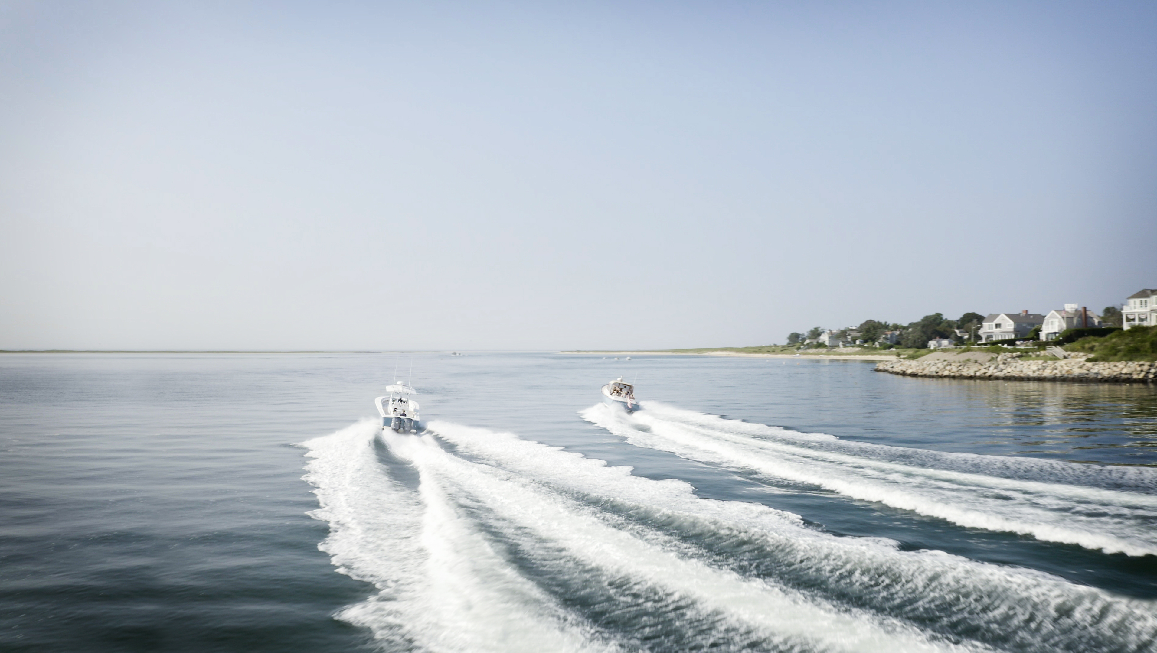 Two motorboats speed across the water leaving white wakes, with houses visible along the shoreline under a clear sky.