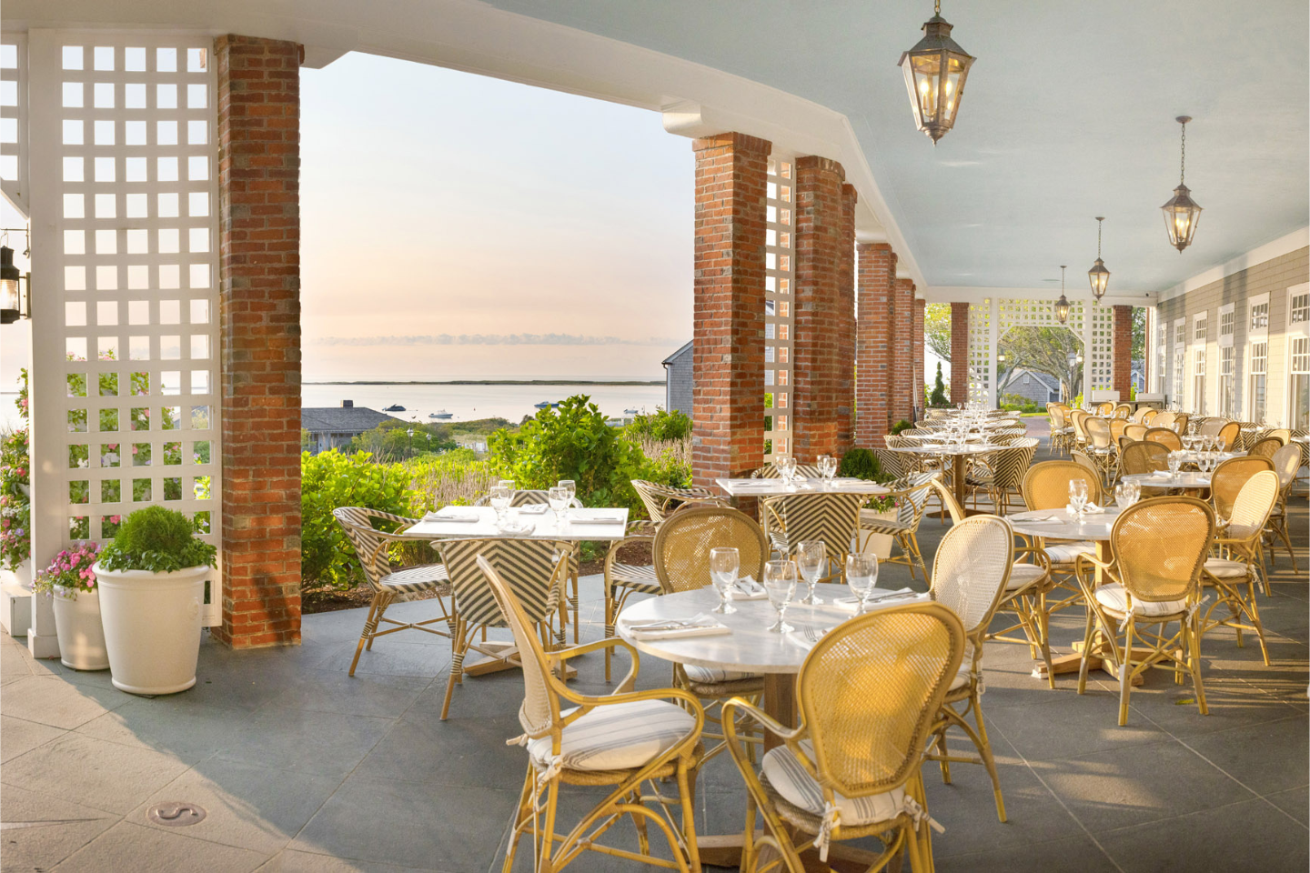 Outdoor restaurant terrace with wicker chairs and tables set for dining, overlooking a scenic view of the sea at sunset.