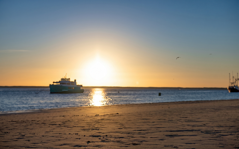 A sandy beach at sunset with a boat on the water and a clear sky.