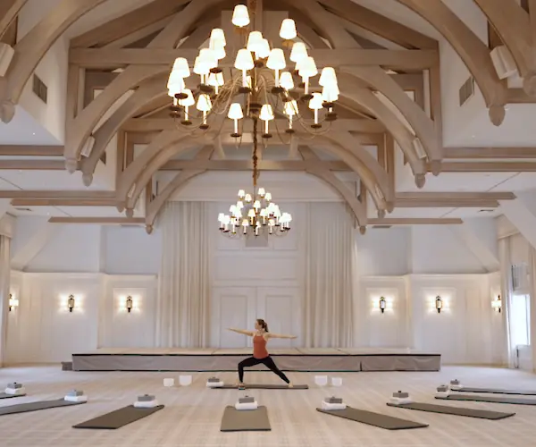 A person is doing yoga in an elegant room with chandeliers and yoga mats arranged in a semicircle on the floor.