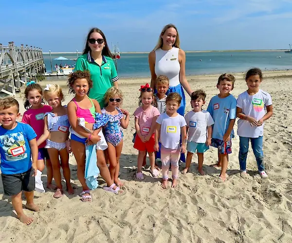 A group of children and two women pose on a sandy beach near a wooden dock, with the ocean and boats in the background.