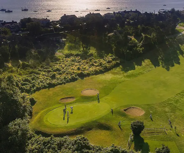Aerial view of a golf course with players, near a coastline with boats and houses, surrounded by lush greenery.