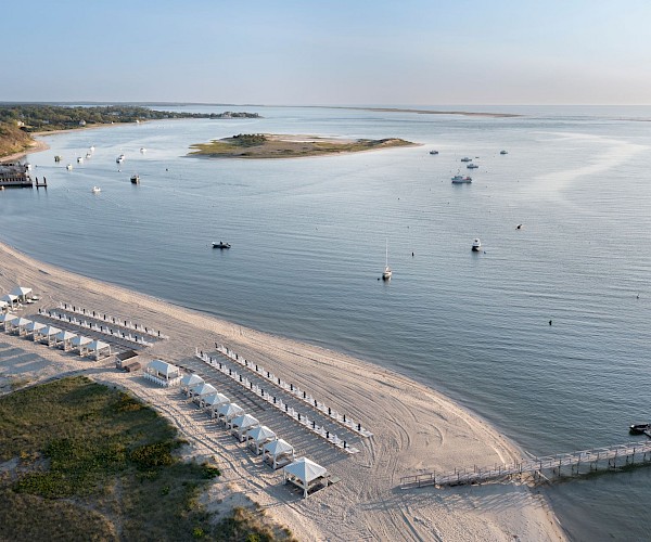 Aerial view of a serene beach with rows of cabanas, a dock, and boats on calm water under a clear sky.