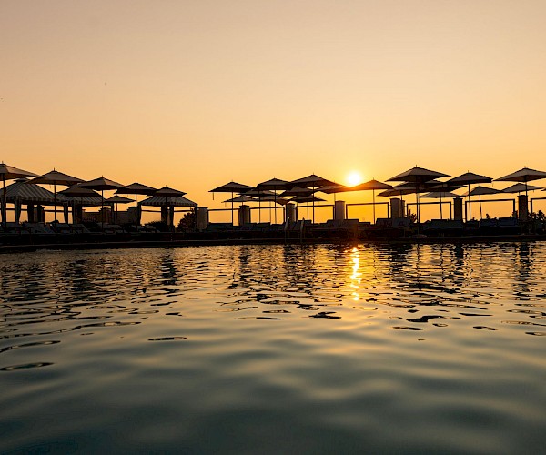 The image shows a serene sunset over a pool, with silhouettes of umbrellas and loungers reflecting on the water's surface.