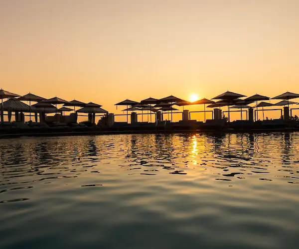 The image shows a serene sunset over a pool, with silhouettes of umbrellas and loungers reflecting on the water's surface.