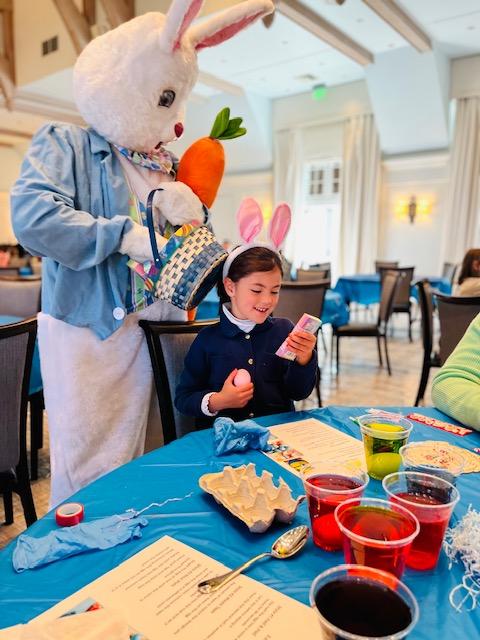 Child with bunny ears holds eggs next to an Easter Bunny in a dining room.