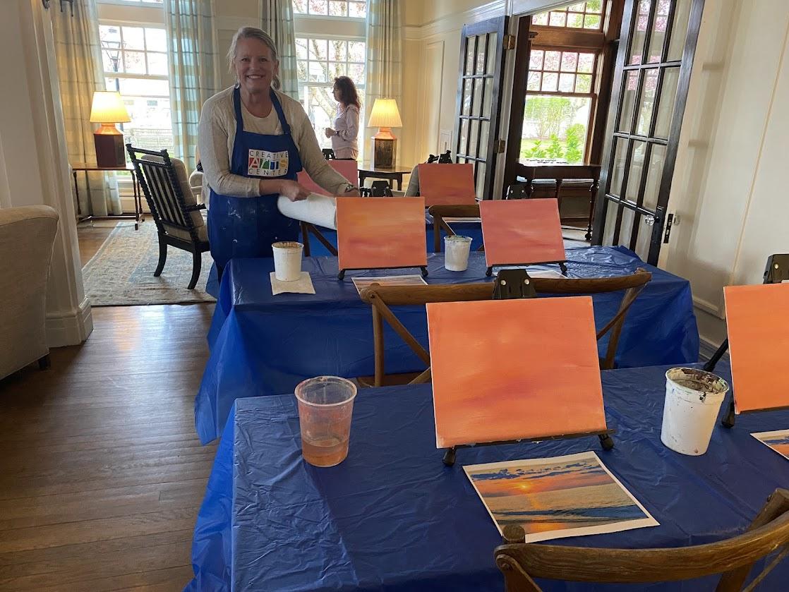 Woman in apron, smiling, standing by tables with painting setups in a sunlit room.