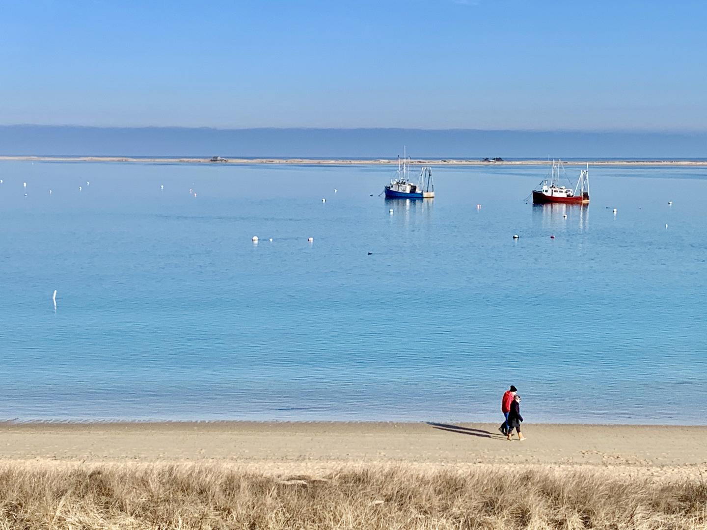 Calm beach, clear blue water, two boats in distance, person walking on sand.