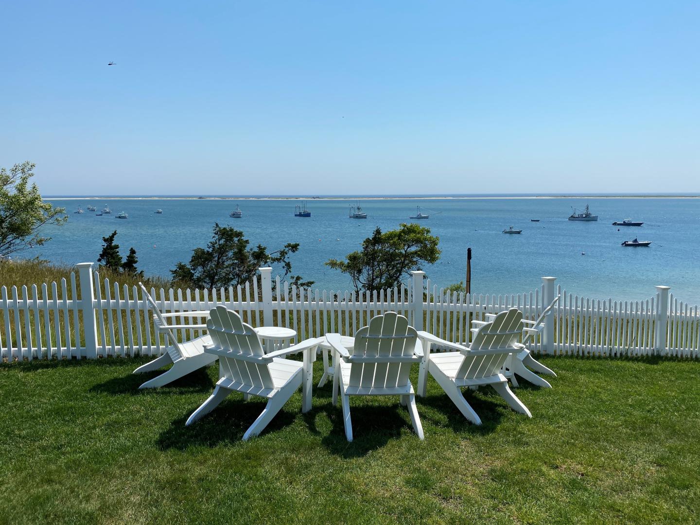 White Adirondack chairs on grass overlook a calm ocean, framed by a white fence.
