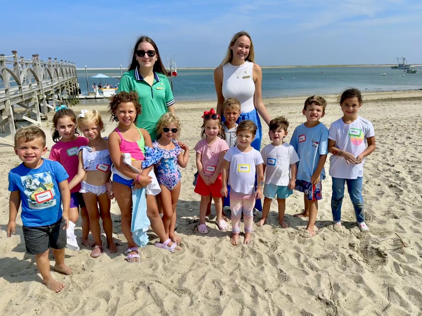 Children with two adults smiling on a sunny beach, with the ocean and pier in the background.