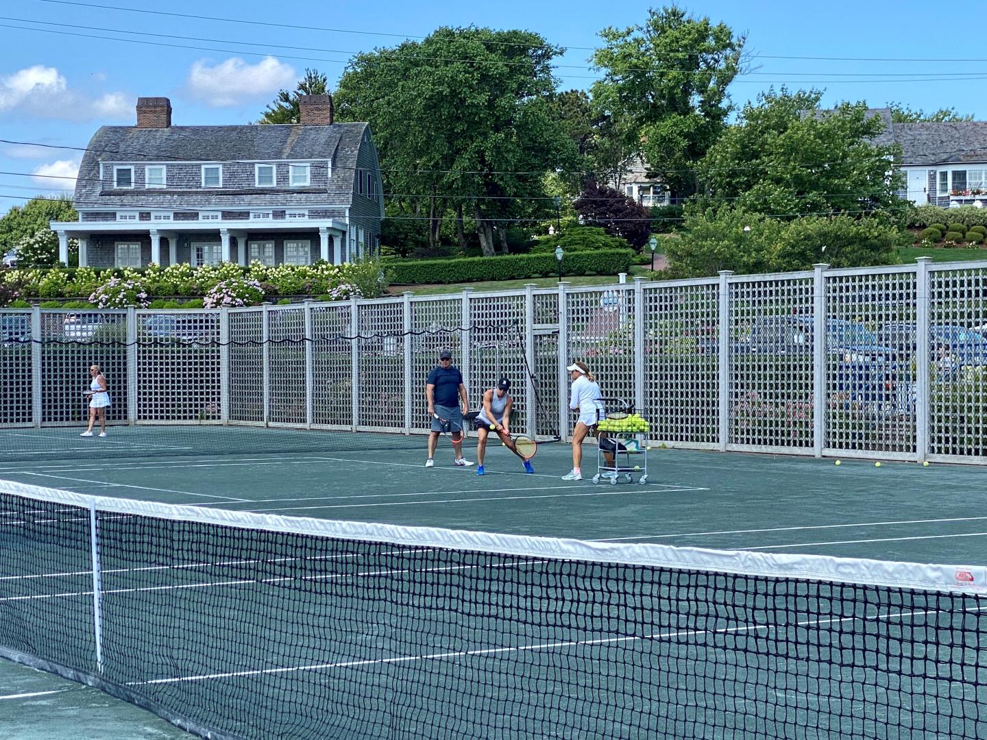 Tennis players practicing on a court with houses and trees in the background.