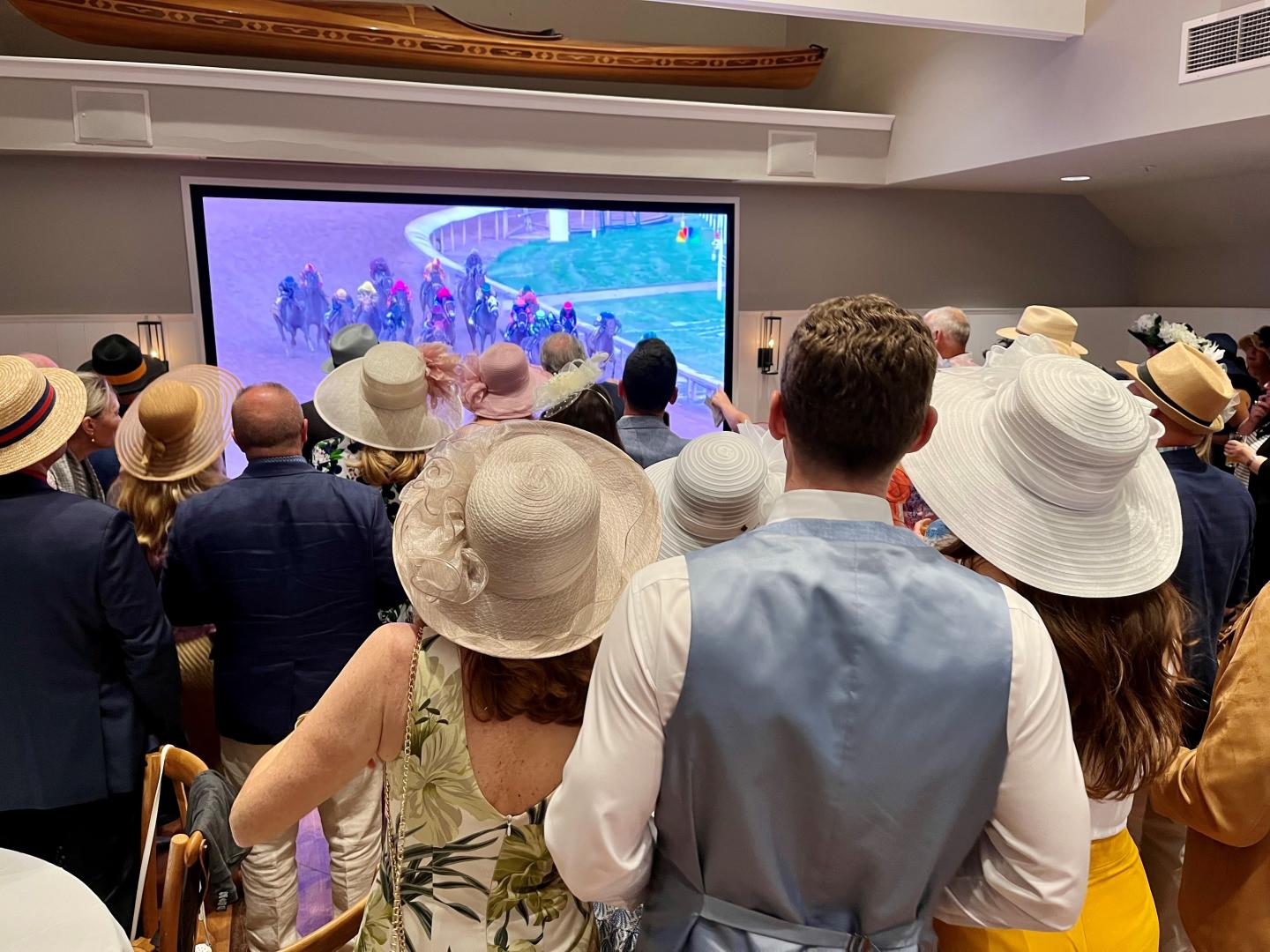People in hats watching a horse race on a large screen indoors.