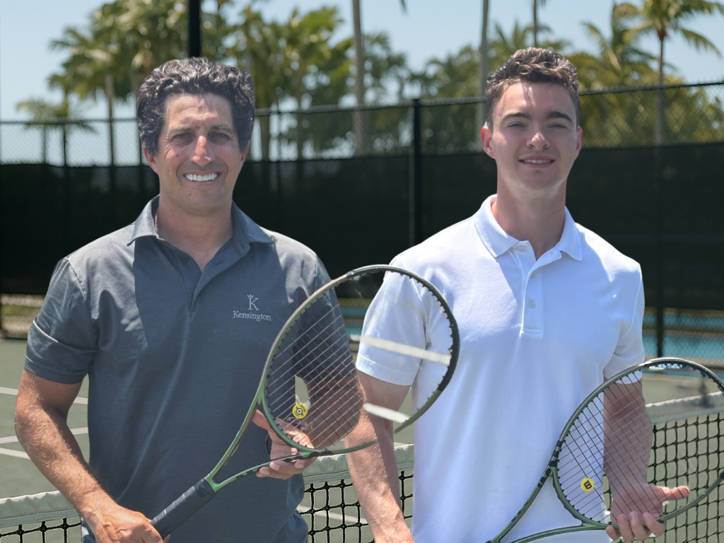 Two men smiling with tennis rackets on a court, palm trees in the background.