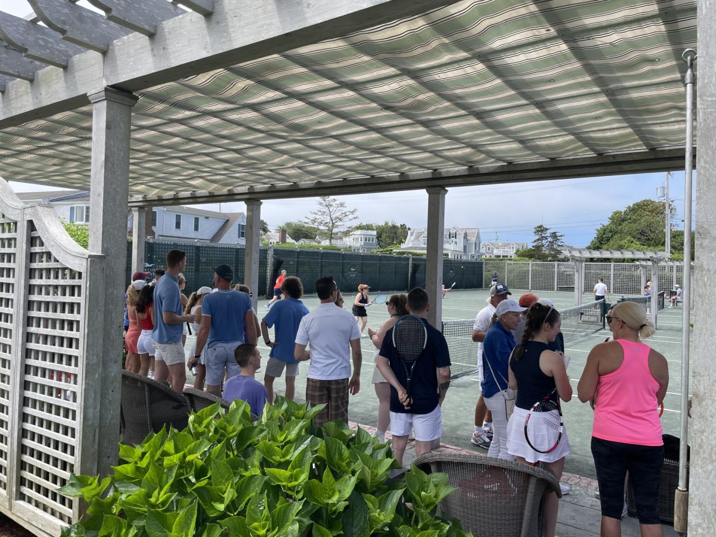 People gather under a pergola near a tennis court on a sunny day.