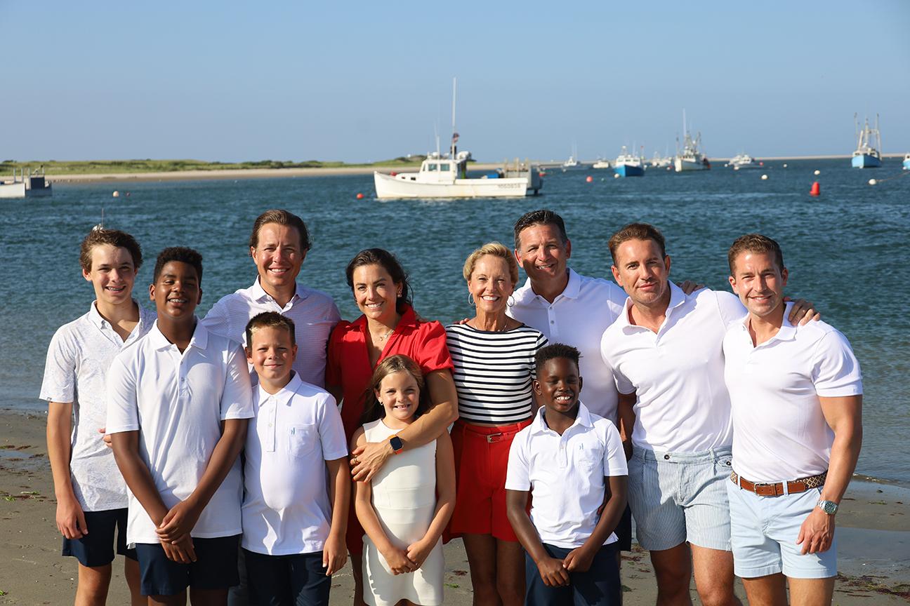 Family smiling on a beach, boats in background on a sunny day.