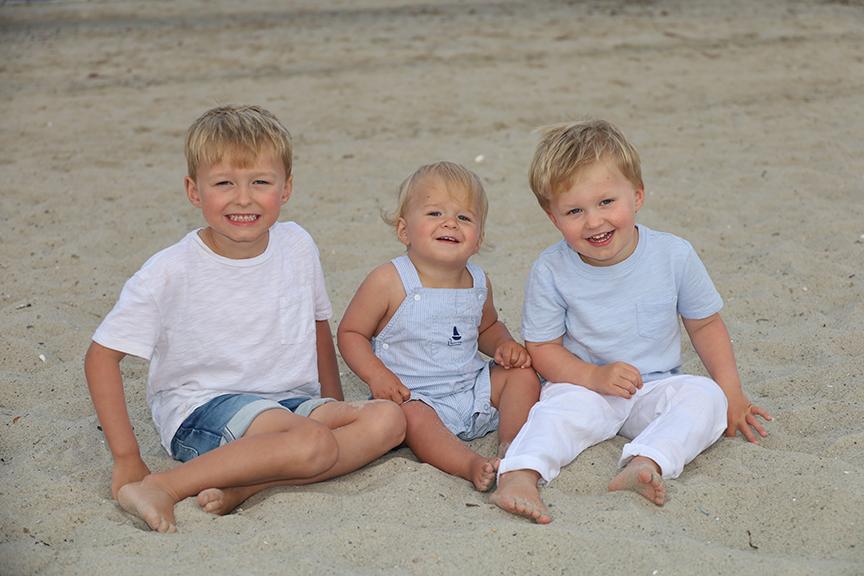 Three smiling children sitting on a sandy beach.