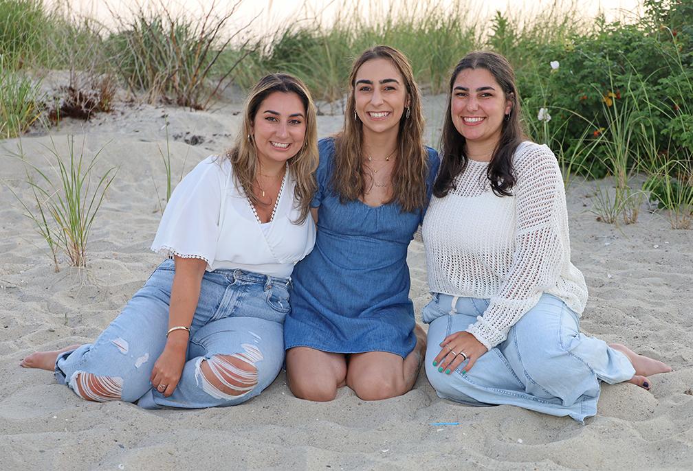 Three people sitting on sand, smiling, with greenery in the background.