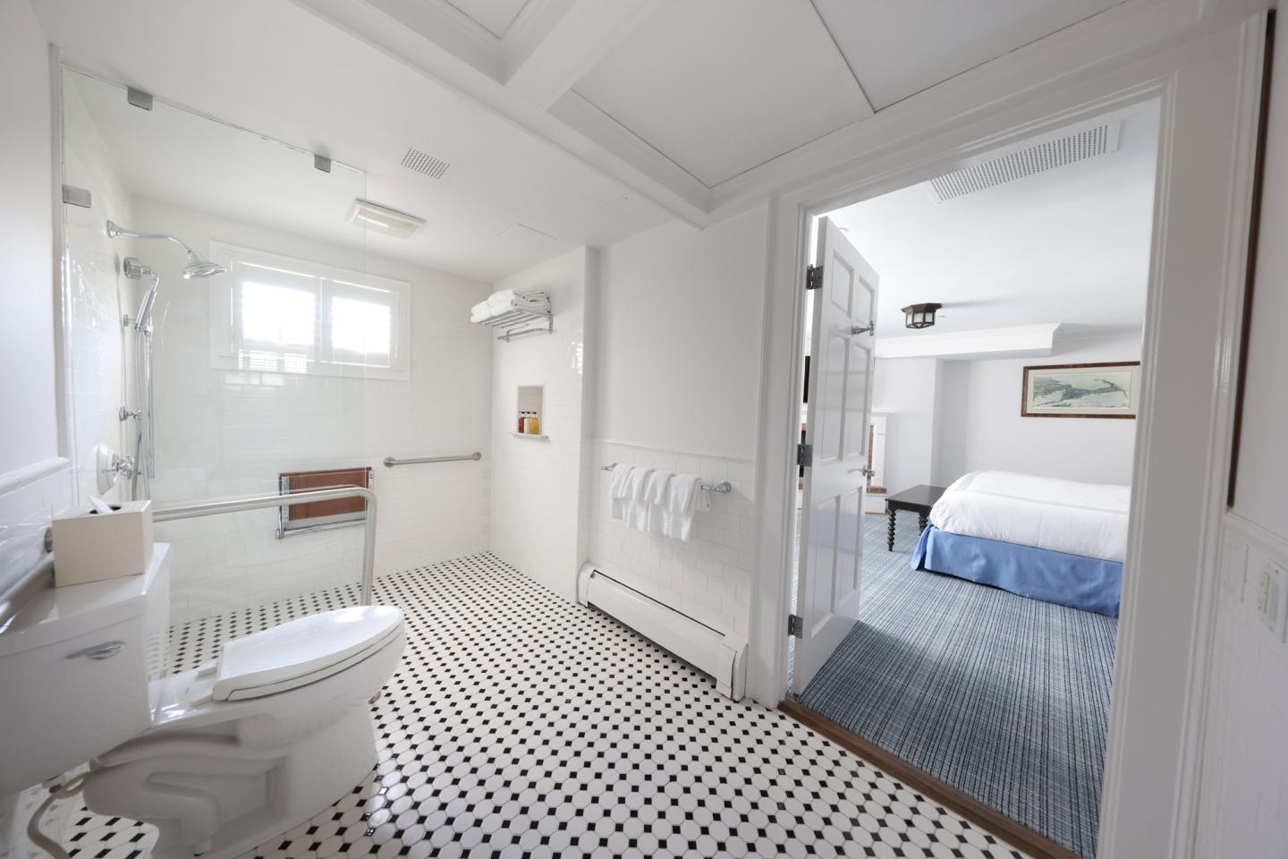 Accessible bathroom with a shower seat, leading to a bedroom with a blue bedspread.