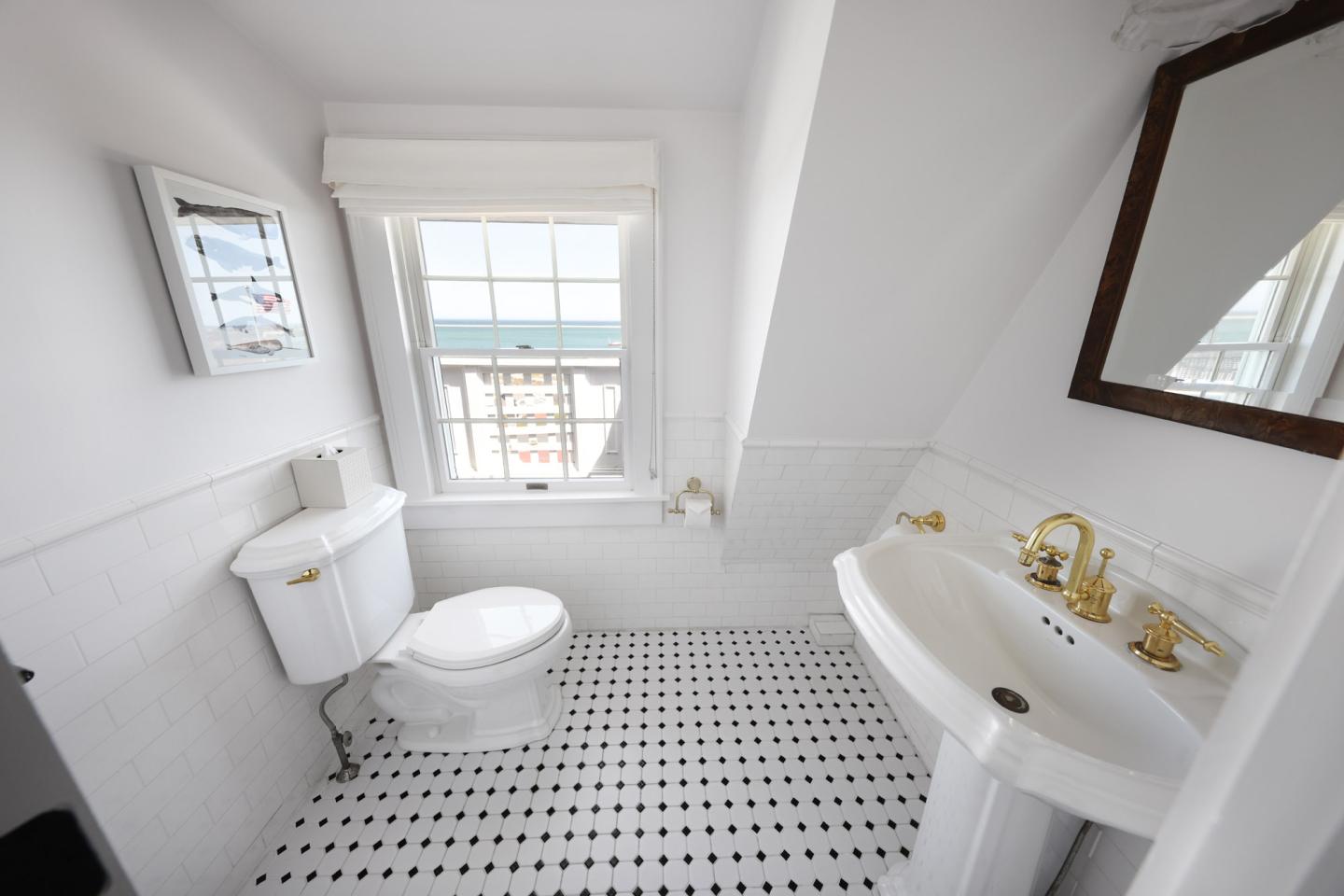 Bright bathroom with white tiles, gold fixtures, and a window with an ocean view.