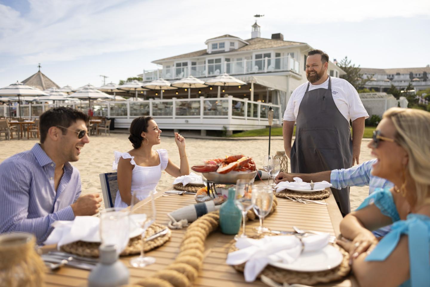 Outdoor dining scene with friends and a chef, seated by a beachside restaurant.