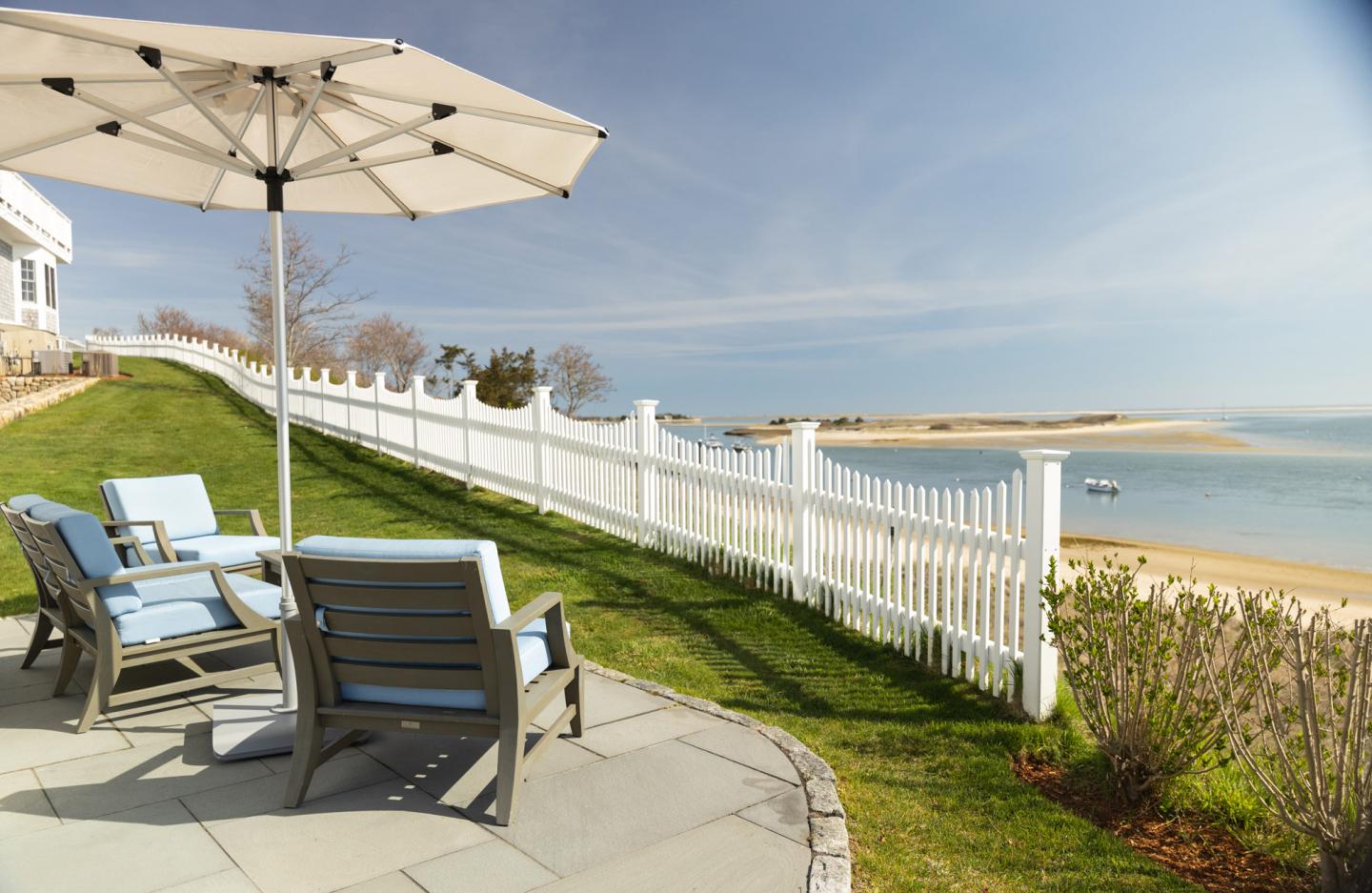 Patio chairs under an umbrella, overlooking a beach and ocean.
