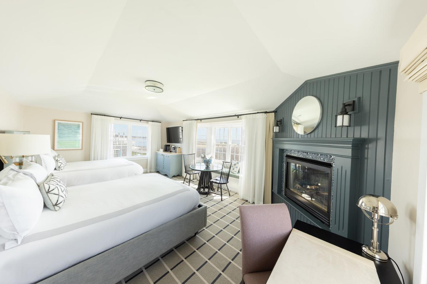 Bright hotel room with two beds, fireplace, and large windows.